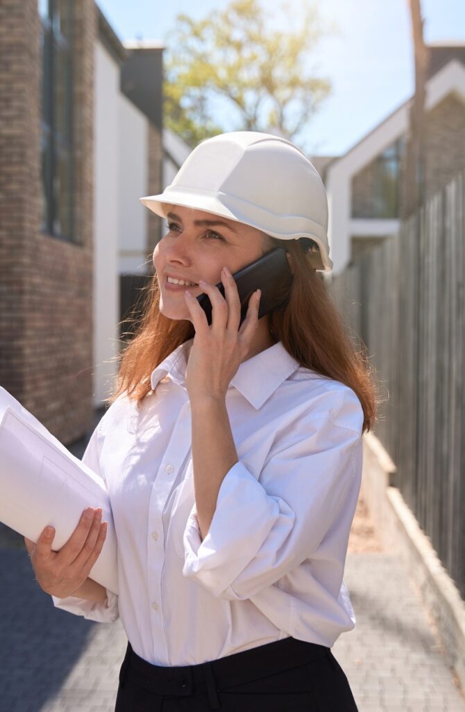 Woman cadastral worker in hardhat holding layouts and talking to mobile phone